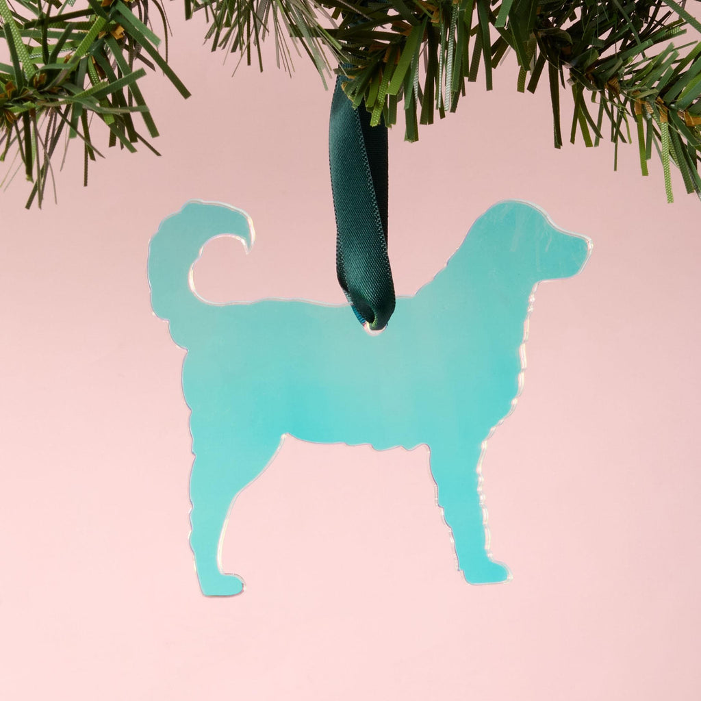 A laser-cut fluffy dog silhouette ornament (such as a Golden Retriever or Spaniel) made from iridescent teal/blue acrylic. The ornament is hanging by a dark green ribbon from a pine branch, set against a pink background.