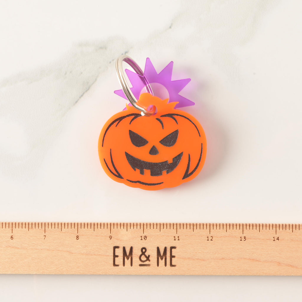 A close-up product photo of an orange jack-o'-lantern shaped pet ID tag with a carved, spooky face. A purple starburst charm is attached to the top of the tag, which is shown with a silver split key ring. A wooden ruler with the text "EM & ME" is positioned below the tag to show its size.