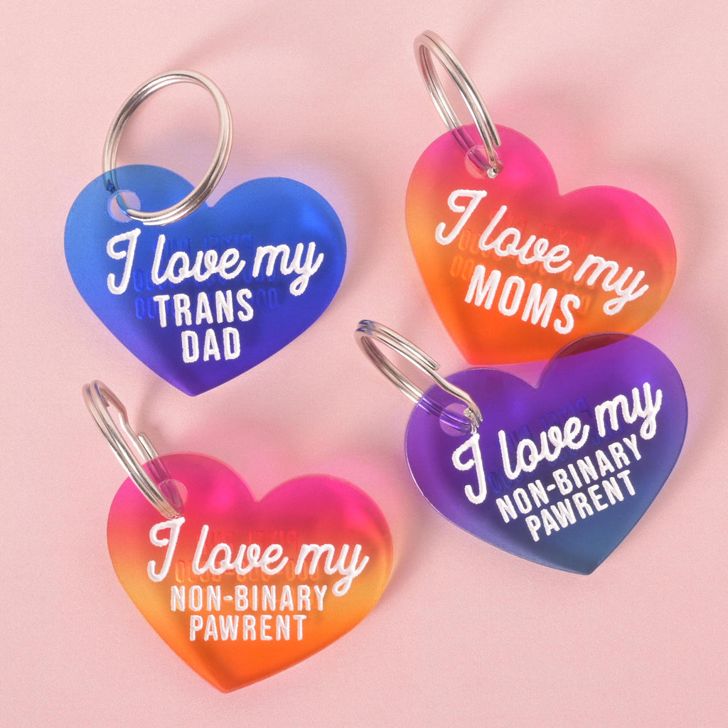 Set of four colorful acrylic heart-shaped pet ID tags with white lettering. The tags read: “I love my trans dad,” “I love my moms,” and “I love my non-binary pawrent” in rainbow and blue gradient designs.