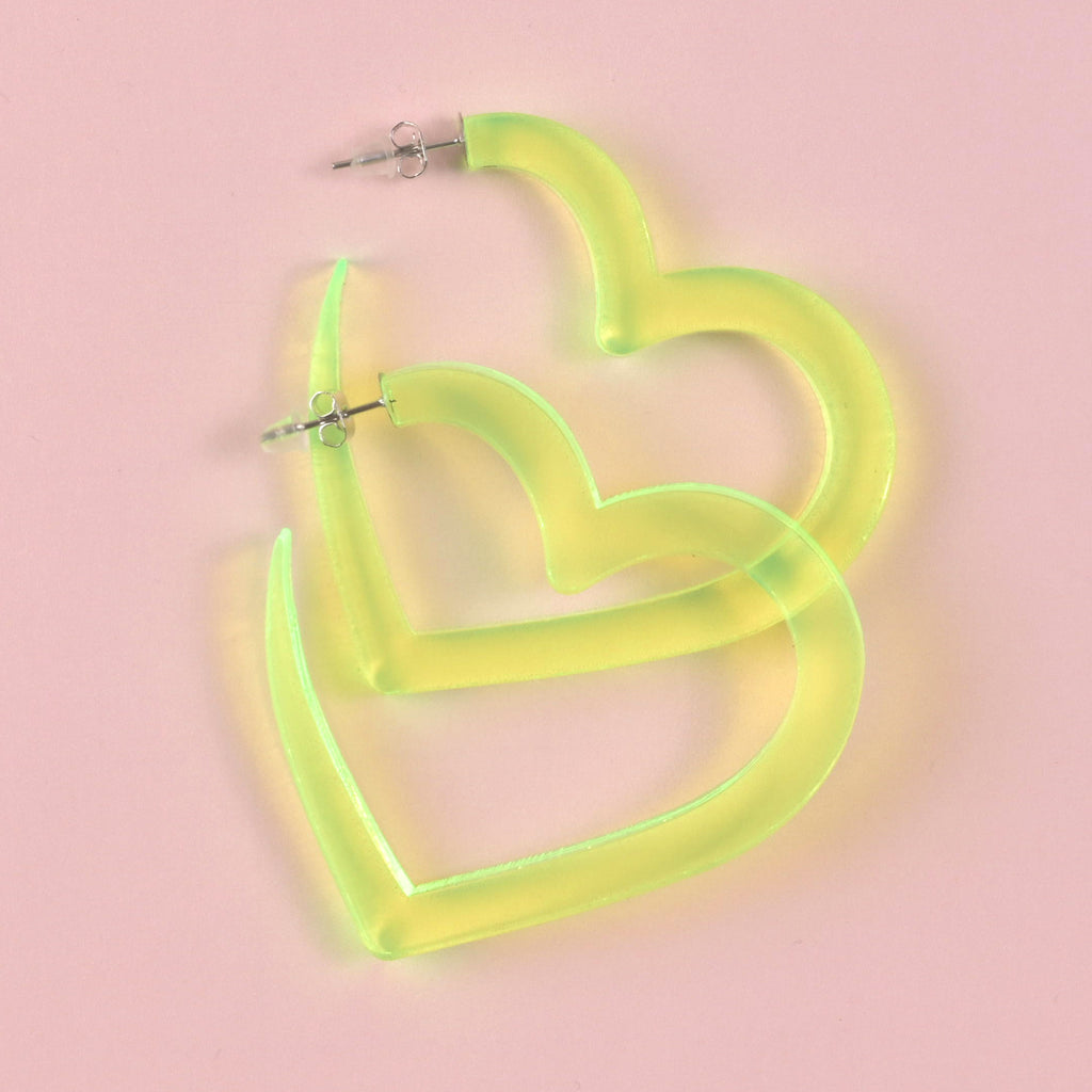 A pair of oversized, fluorescent neon yellow-green acrylic heart-shaped hoop earrings with silver posts, displayed on a solid pink background.