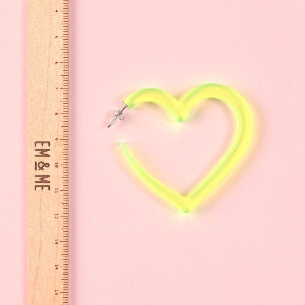 A single oversized fluorescent neon yellow-green acrylic heart hoop earring shown next to a wooden ruler marked with the 'EM & ME' logo to clearly indicate its large size.