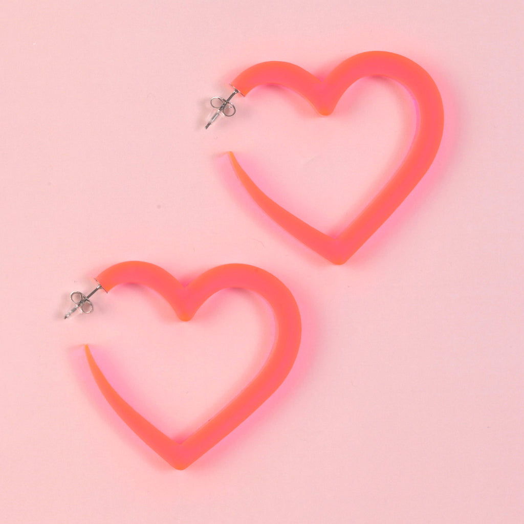 A pair of oversized, fluorescent coral/pink acrylic heart-shaped hoop earrings with silver posts, displayed on a solid pink background.