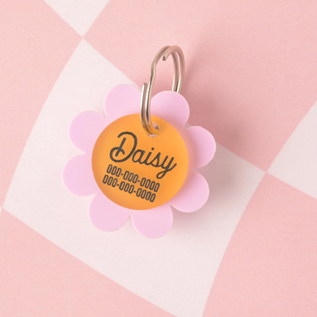 Acrylic pet ID tag shaped like a daisy with light purple (lavender) petals and a yellow center, engraved with the name Daisy and two contact phone numbers, shown on a pink checkered background