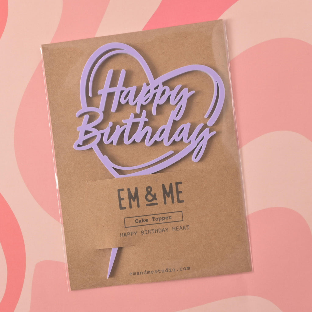 A lavender cake topper that says "Happy Birthday" inside a heart outline, sealed in its retail packaging. The brown cardstock packaging has "EM & ME" printed on it. The item is shown on a pink background with a wavy design.