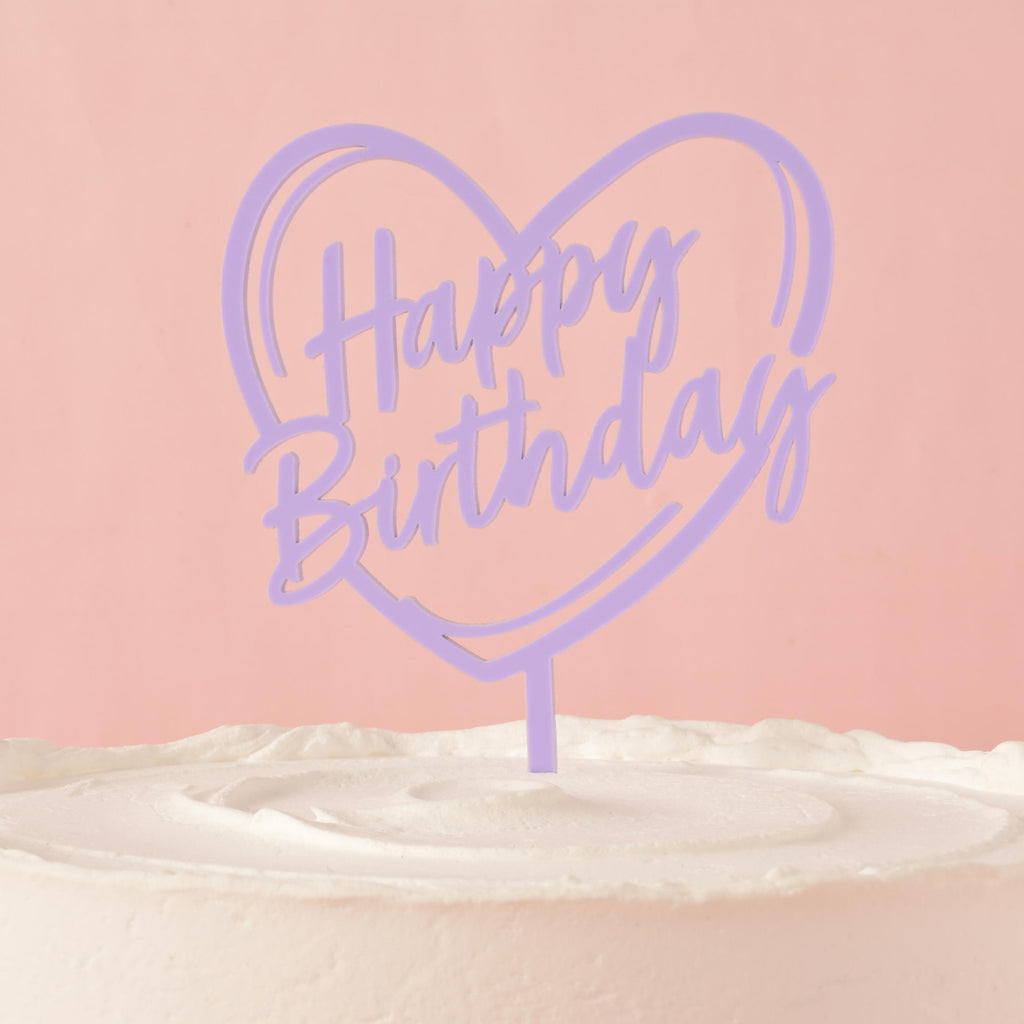 A lavender cake topper that says "Happy Birthday" inside a heart outline, placed on top of a frosted white cake. The background is a solid pink color.
