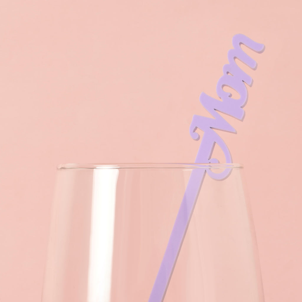 A single light purple cocktail stirrer with the word "Mom" is placed inside a clear glass. The background is a solid pink color.