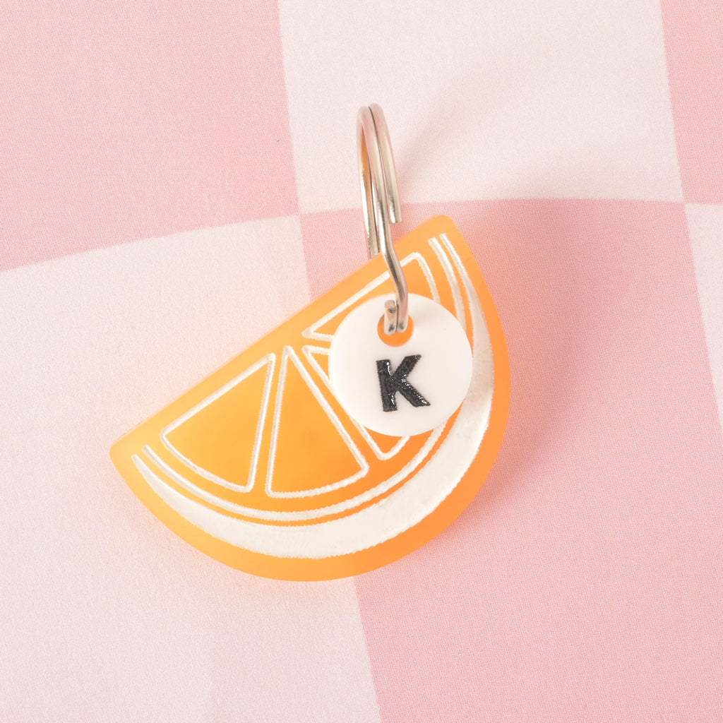 A custom, translucent yellow, lemo slice-shaped pet ID tag with a white outline. A small, white, circular charm with a black letter "K" is attached to the silver keyring. Both charms are displayed on a pink and white checkered background.