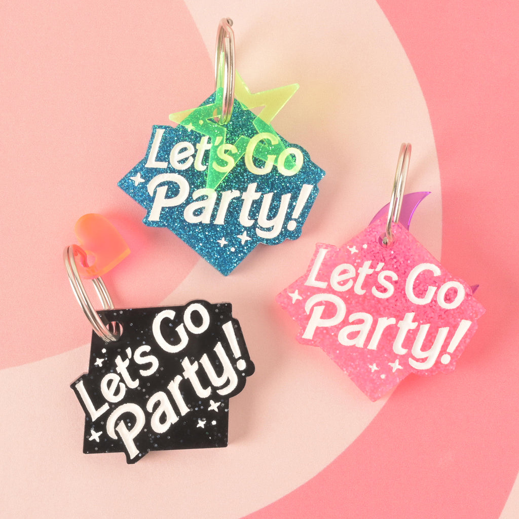 A product photo showing three glittery, rhombus-shaped pet ID tags. The tag on the top is blue with a green star charm. The tag on the bottom left is black with a neon orange heart charm. The tag on the bottom right is pink with a purple geometric charm. All three tags are engraved with the words "Let's Go Party!" and have a silver split key ring.