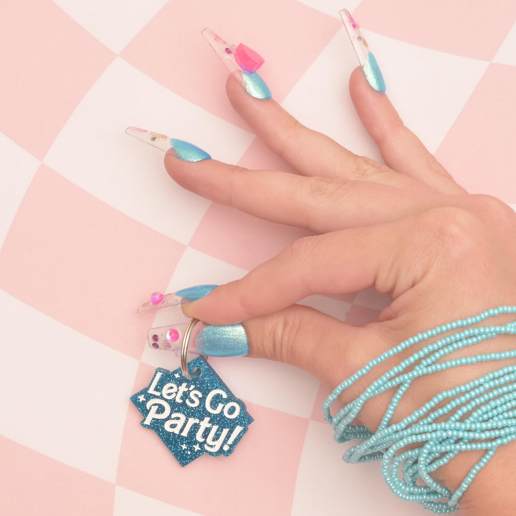 Close up of hands with blue nails holding a blue glitter Barbie inspired pet tag that reads "Let's go party!" on pick checked background