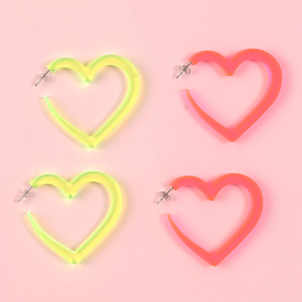 Four large, acrylic heart hoop earrings: two in vibrant neon yellow-green and two in fluorescent pink/orange. The statement earrings are displayed on a solid pink background, highlighting the bold color contrast.