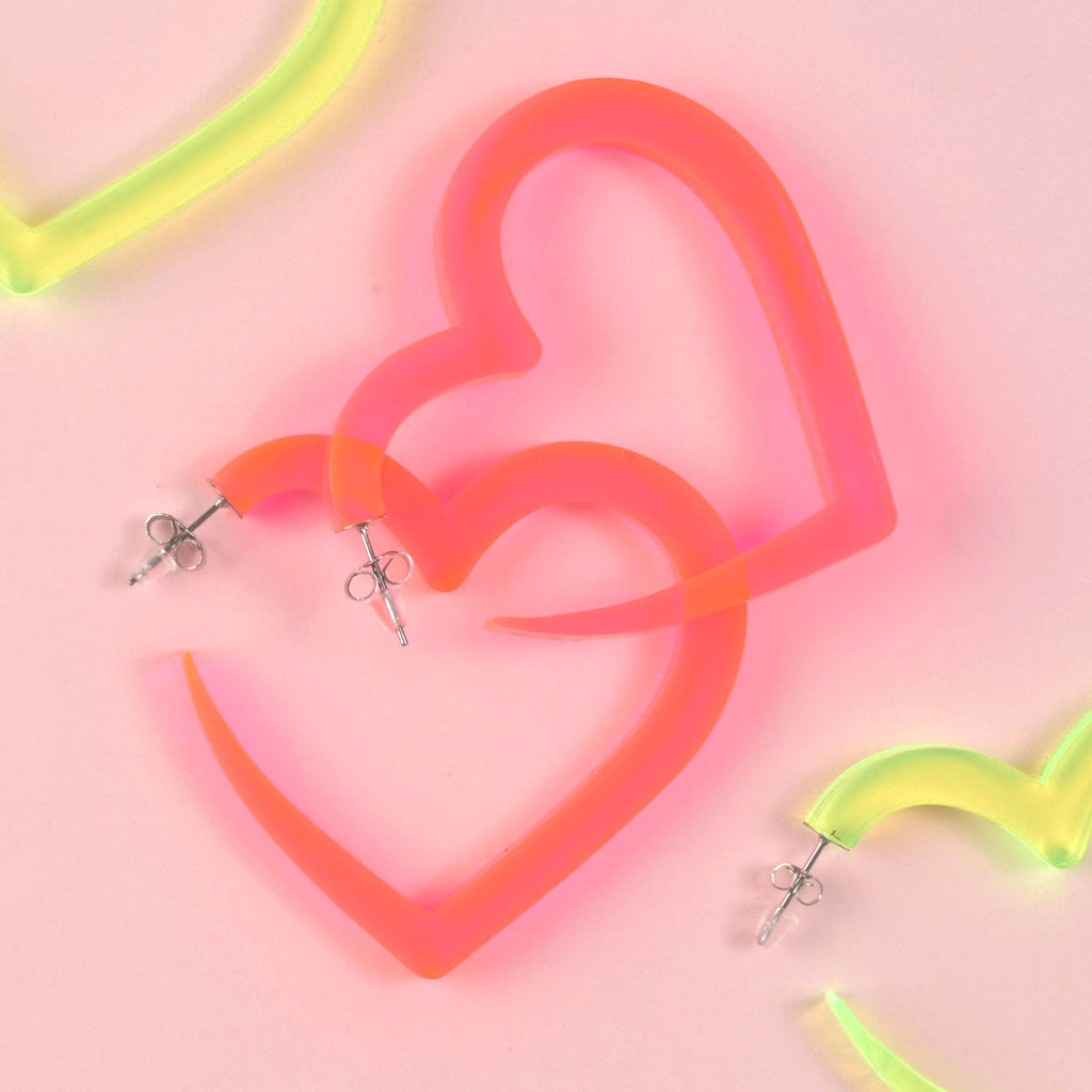 A pair of oversized, fluorescent pink acrylic heart-shaped hoop earrings with silver posts, shown overlapping on a pink background. Hints of a neon yellow pair are visible at the edges.