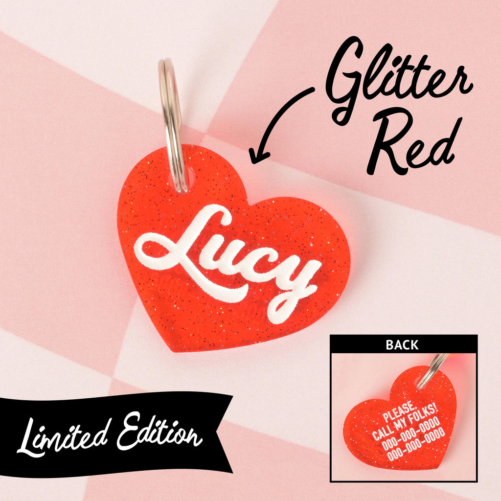 Limited edition glitter red plastic heart-shaped pet ID tag with the name Lucy in white script, shown on a pink checkered background; inset image displays the back of the tag with engraved contact info and the phrase 'Please, call my folks!'