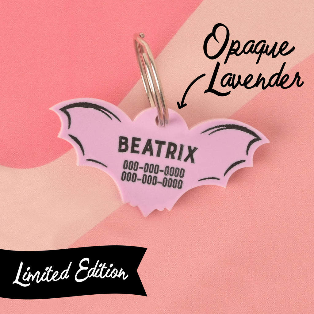 A close-up shot of a custom, limited edition, light purple bat-shaped pet ID tag. The tag is engraved with the name "BEATRIX" and a phone number in black font. The wings have a black, scalloped outline. The tag is attached to a silver keyring. Text on the image labels it "Opaque Lavender" and "Limited Edition." The background is a pastel pink and cream color-blocked pattern.