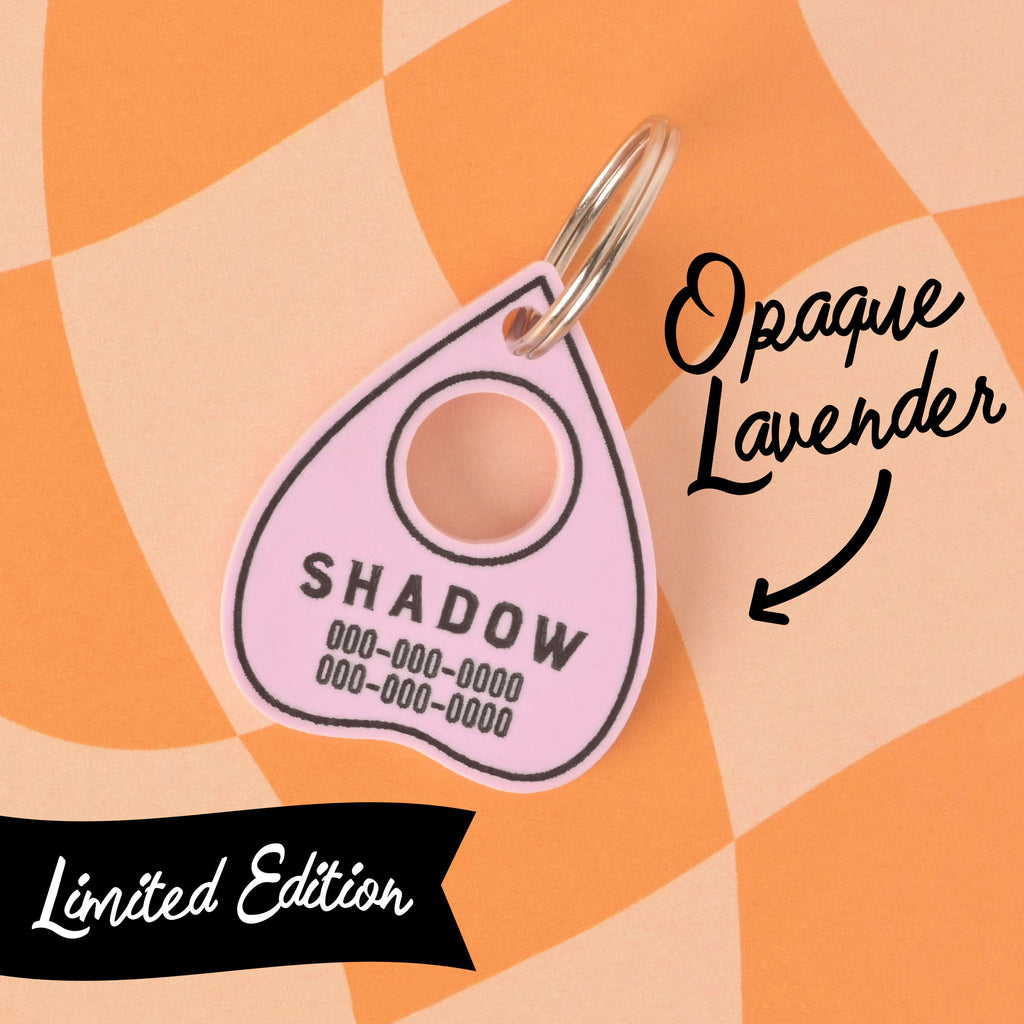 A close-up shot of a custom, limited edition, opaque lavender planchette-shaped pet ID tag. The tag has a black outline and is engraved with the name "SHADOW" and a phone number in black font. The tag is attached to a silver keyring. Text on the image points to the tag, labeling it "Opaque Lavender." A "Limited Edition" ribbon is in the bottom left corner. The tag is shown on a checkered orange and beige background.