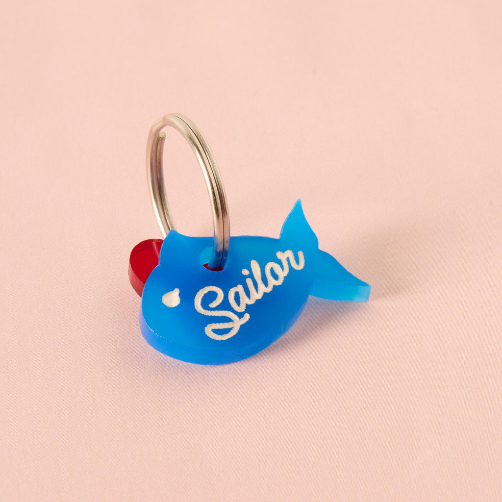 This pet tag is shaped like a blue fish, but instead of a bow, it has a small red heart attached near the top. The word written on it is “Sailor” in white cursive script. The keychain is looped onto a metal ring and placed against a pale pink background.