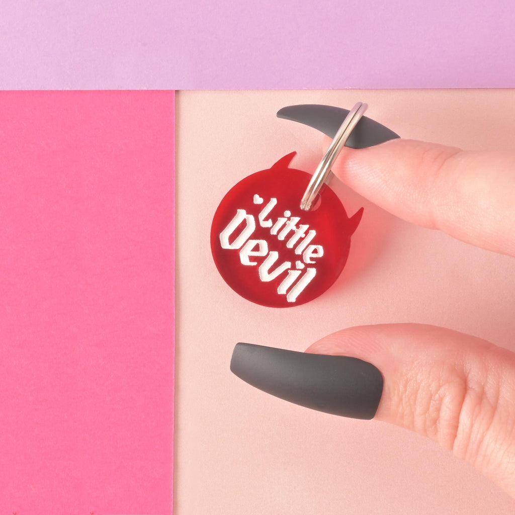 Red little devil personalized pet tag held by a hand with long black fingernails on a pink, purple and peach colored background.