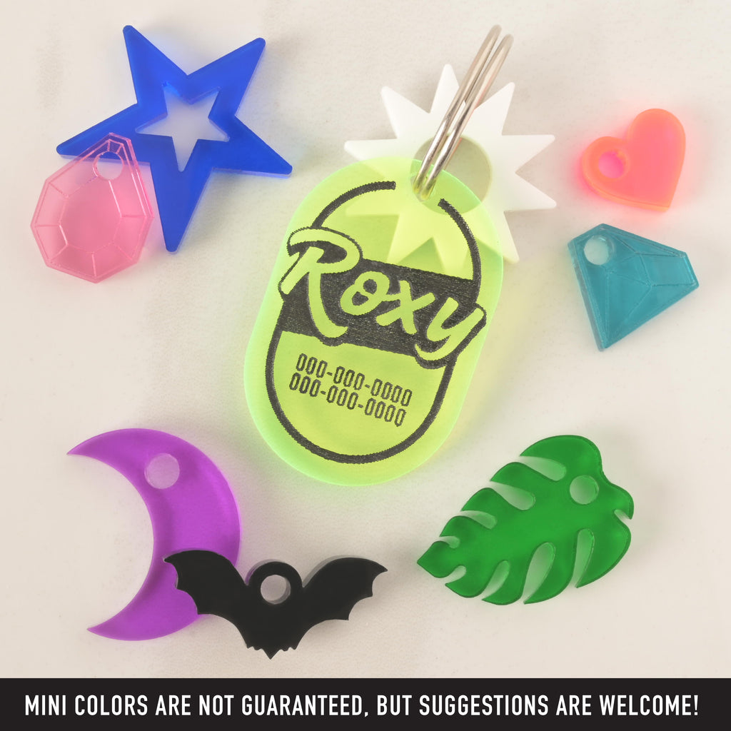 A collection of pet tags and charms scattered on a white surface. The central piece is a personalized neon yellow tag with the name "Roxy" and a placeholder phone number. It has a white starburst charm attached. Surrounding it are various other charms, including a blue star with a pink gemstone, a purple moon, a black bat, a green monstera leaf, a neon pink heart, and a blue diamond. The image also contains the text "MINI COLORS ARE NOT GUARANTEED, BUT SUGGESTIONS ARE WELCOME!"