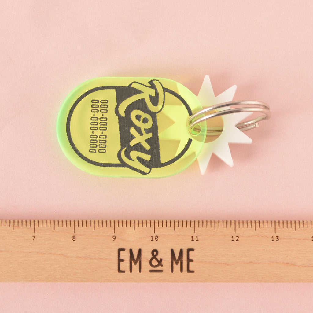 A personalized Fluorescent Green pet tag with a retro, oval shape. The tag has the name "Roxy" and a placeholder phone number on it. A small white starburst charm is attached. A ruler with the "EM & ME" logo is placed below the tag to show its size. The items are shown on a pink background with a wavy design.