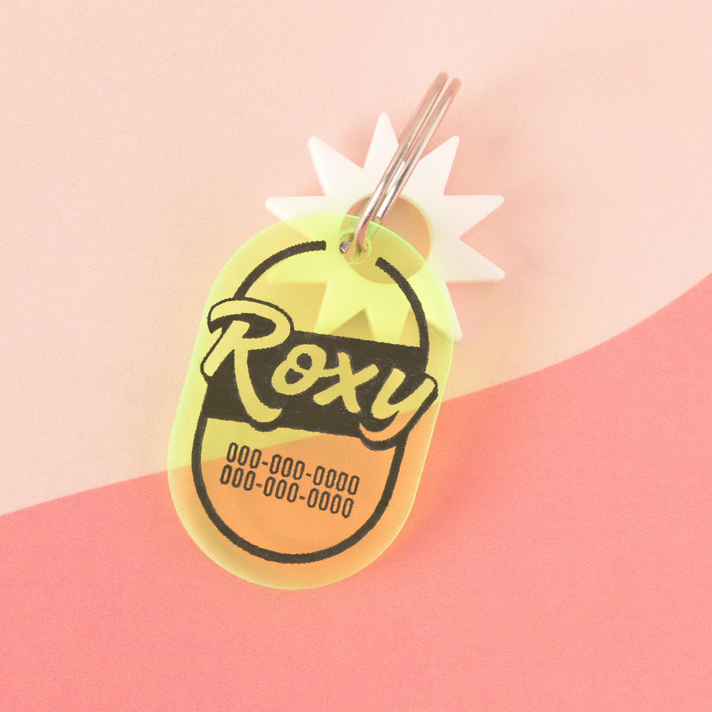 A personalized translucent yellow pet tag with a retro, oval shape. The tag has the name "Roxy" and a placeholder phone number on it. It has a white starburst charm attached and is connected to a silver ring, perfect for a collar. The items are shown on a pink background with a wavy design.