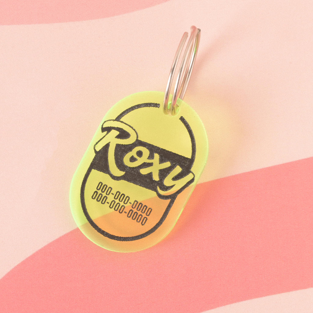 A personalized fluorescent Green pet tag with a retro, oval shape. The tag has the name "Roxy" and a placeholder phone number on it. It's attached to a silver ring, perfect for a collar, and is displayed on a pink background with a wavy design.