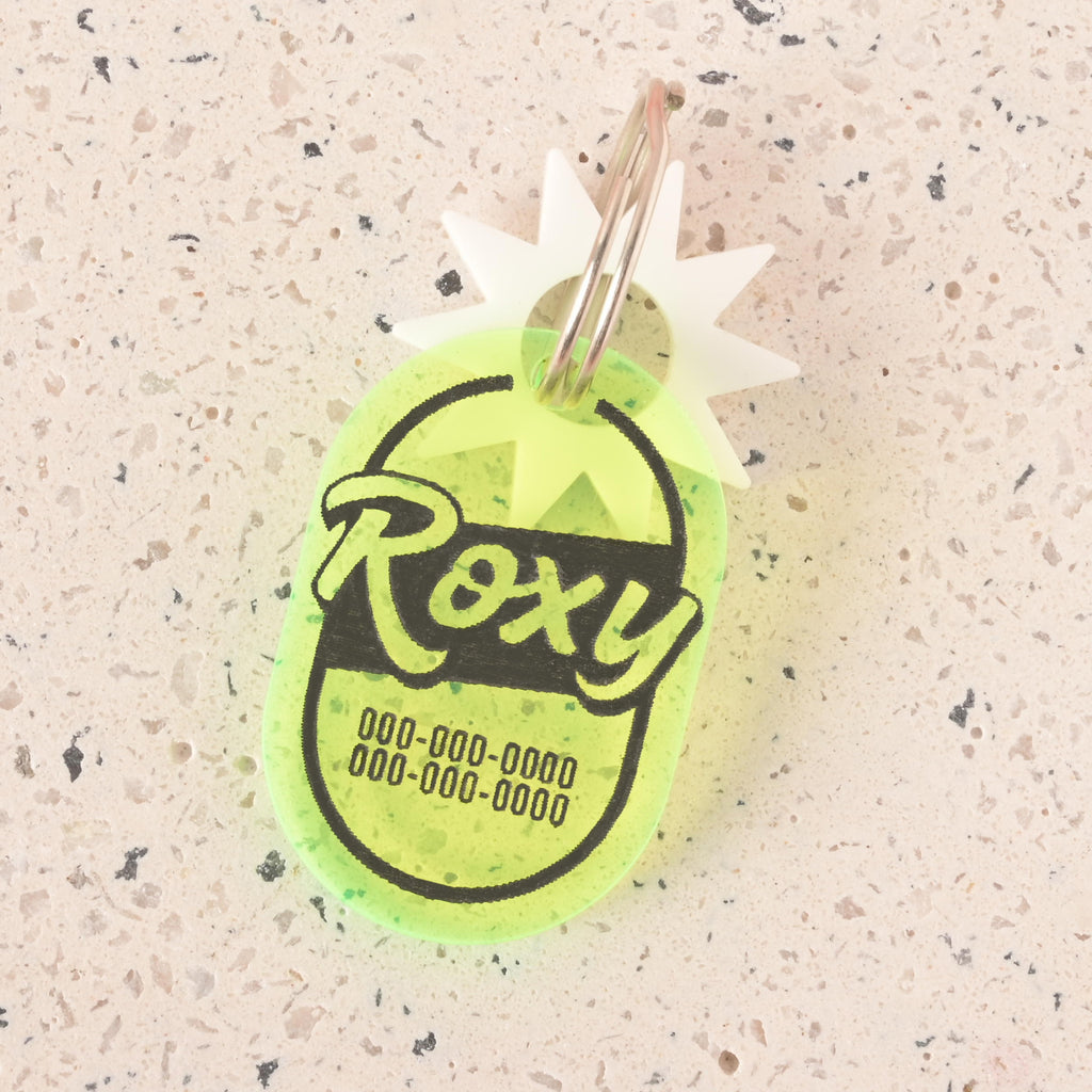 A personalized translucent yellow pet tag with a retro, oval shape. The tag has the name "Roxy" and a placeholder phone number on it. It has a white starburst charm attached and is connected to a silver ring, perfect for a collar. The items are shown on a light-colored, speckled surface.