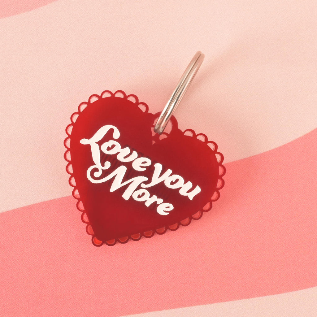 A red heart-shaped pet tag with a wavy edge that says "Love you More" in white letters. The tag is attached to a silver ring, perfect for a collar, and is resting on a pink background with a wavy design.