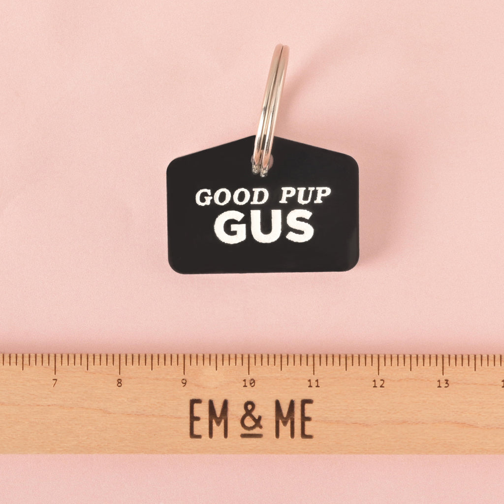 A black dog tag that says "GOOD PUP GUS" in white letters. A ruler with the "EM & ME" logo is placed below the tag to show its size. The tag is attached to a silver ring and is resting on a solid pink background.