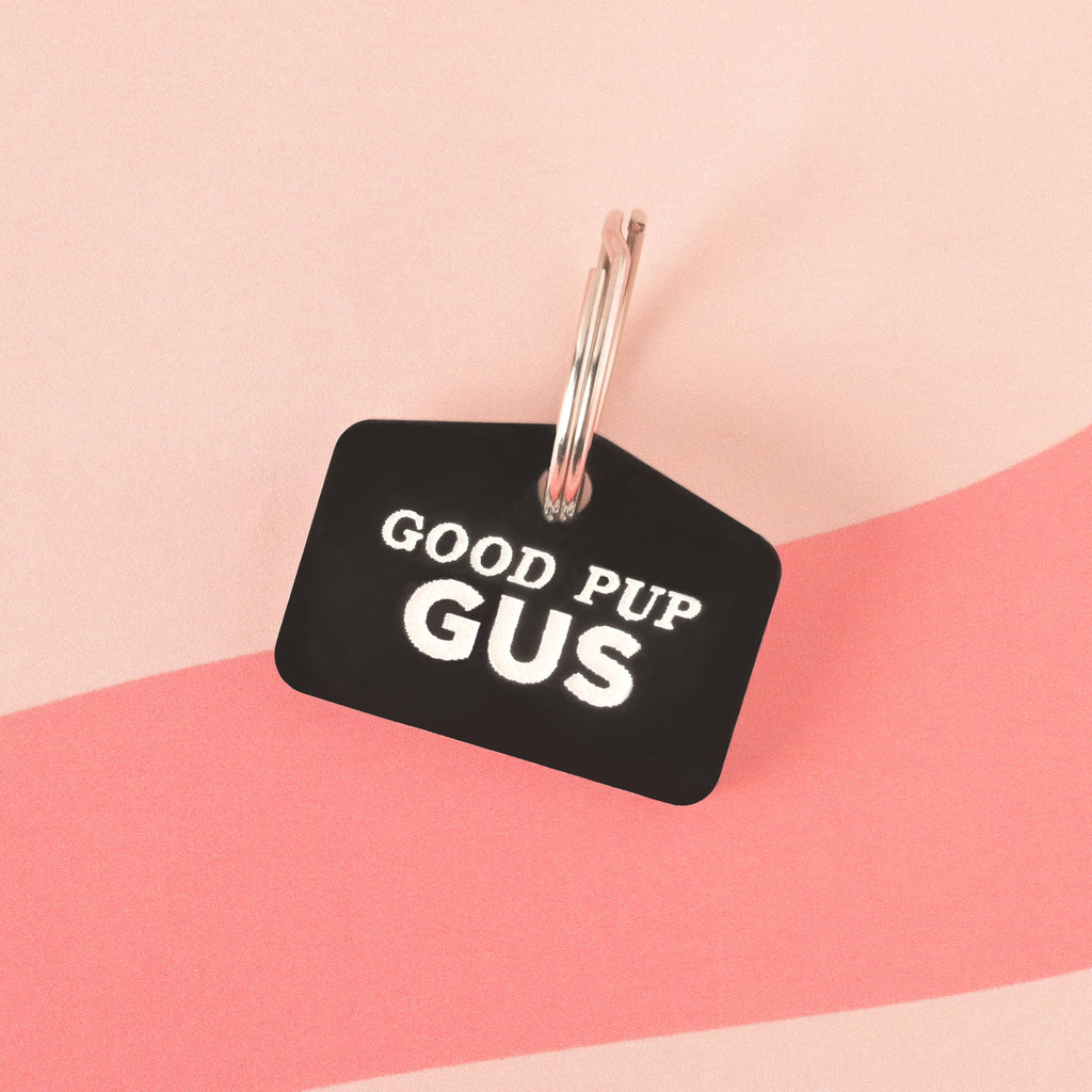 A black dog tag that says "GOOD PUP GUS" in white letters. The tag is attached to a silver ring and is resting on a pink background with a wavy design.