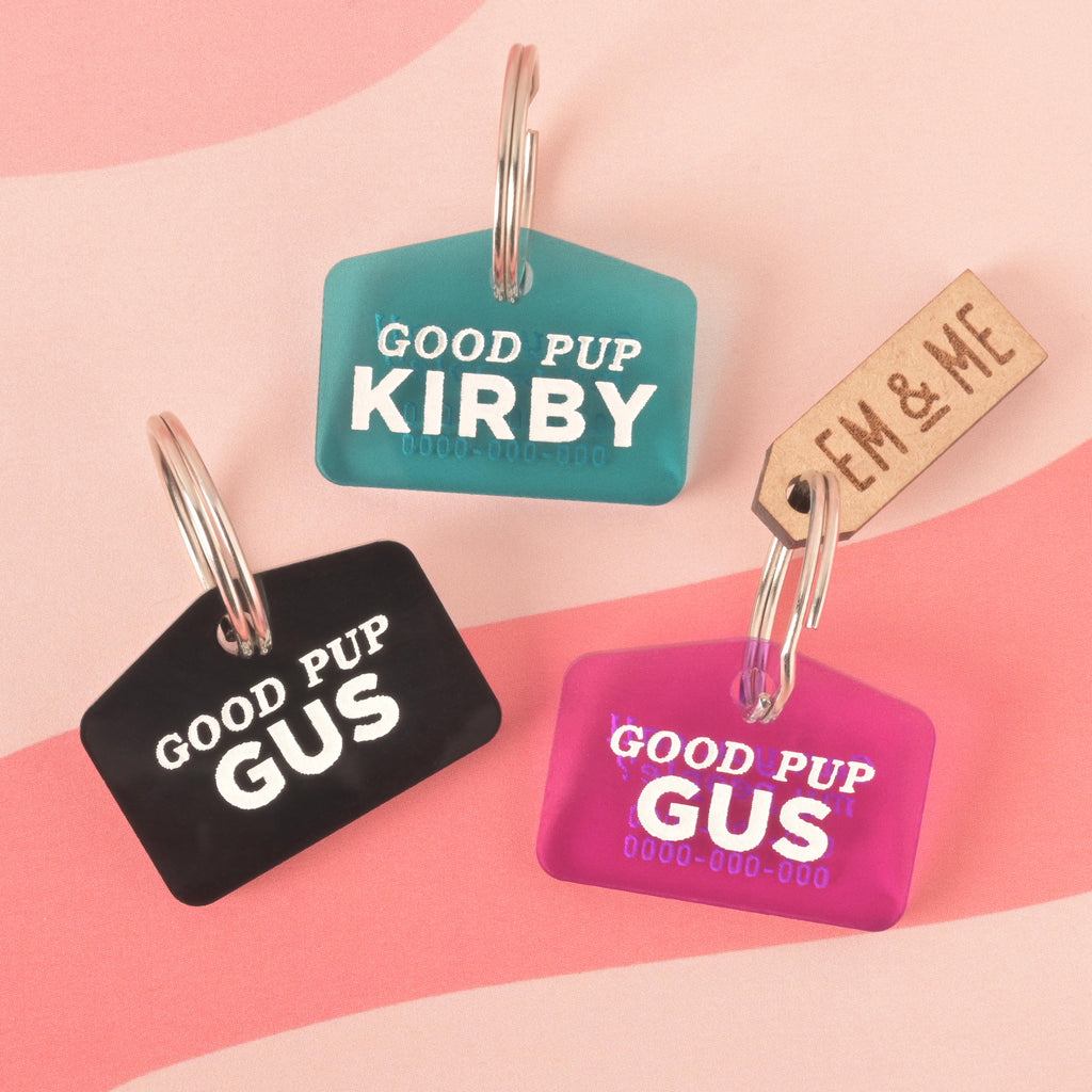 A set of three "Good Pup" dog tags in different colors. One black tag says "Gus," a blue-green tag says "Kirby," and a pink tag says "Gus." All tags are attached to a silver ring for a collar. A small rectangular wooden charm that reads "EM & ME" is also visible. The items are shown on a light pink and red wavy background.