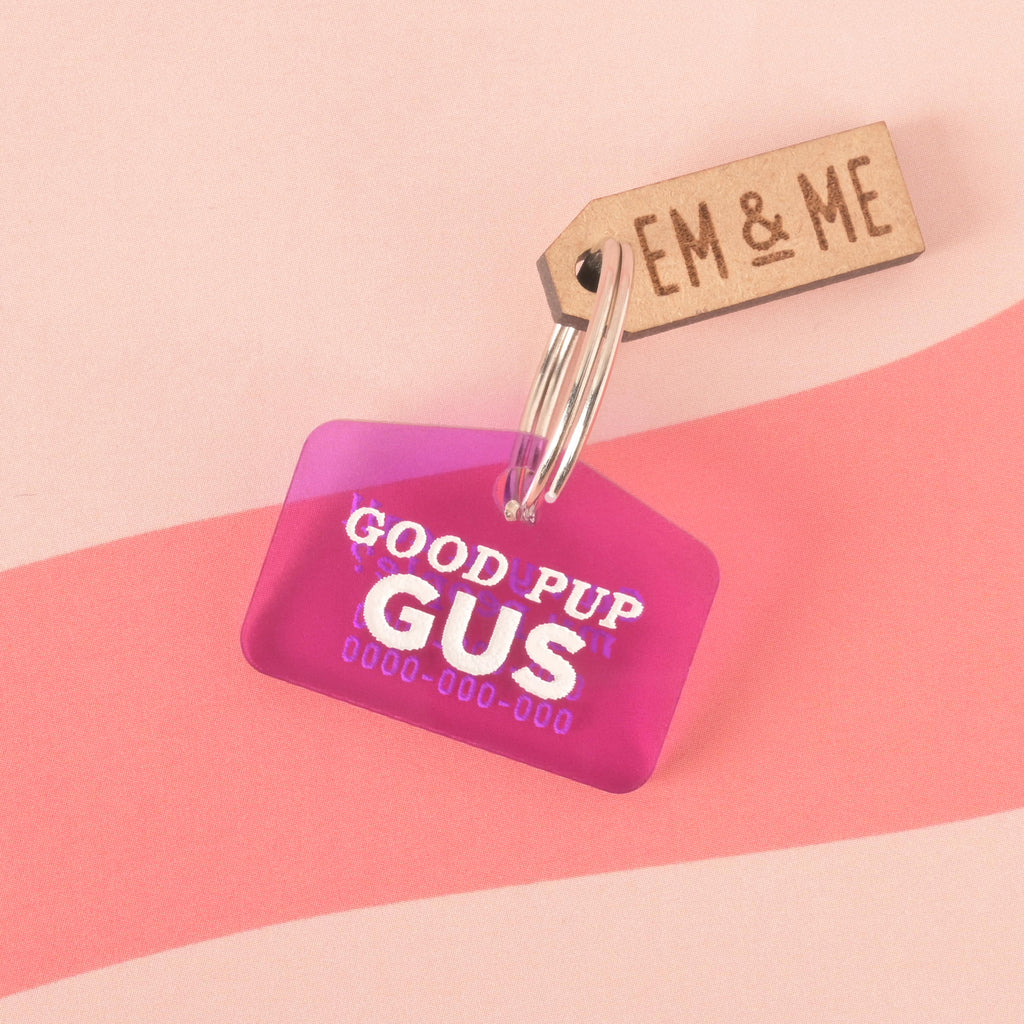 A purple  dog tag that says "GOOD PUP GUS" in white letters with a placeholder phone number. A small wooden charm that says "EM & ME" is attached to the tag. Both are connected to a silver ring and are resting on a pink background with a wavy design.