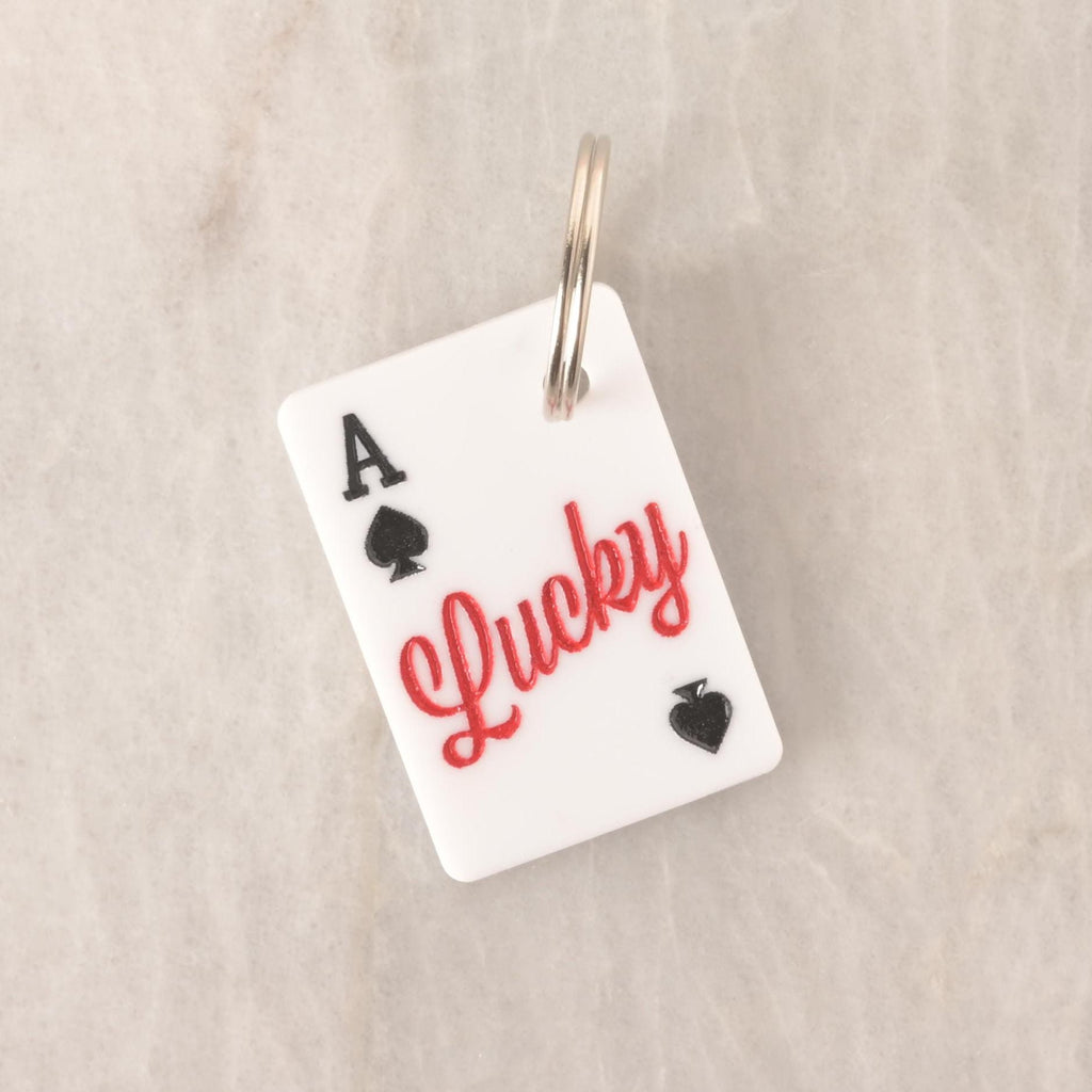 White acrylic Lucky Ace pet ID tag shaped like a playing card with black spades and red “Lucky” text, attached to a silver keyring.