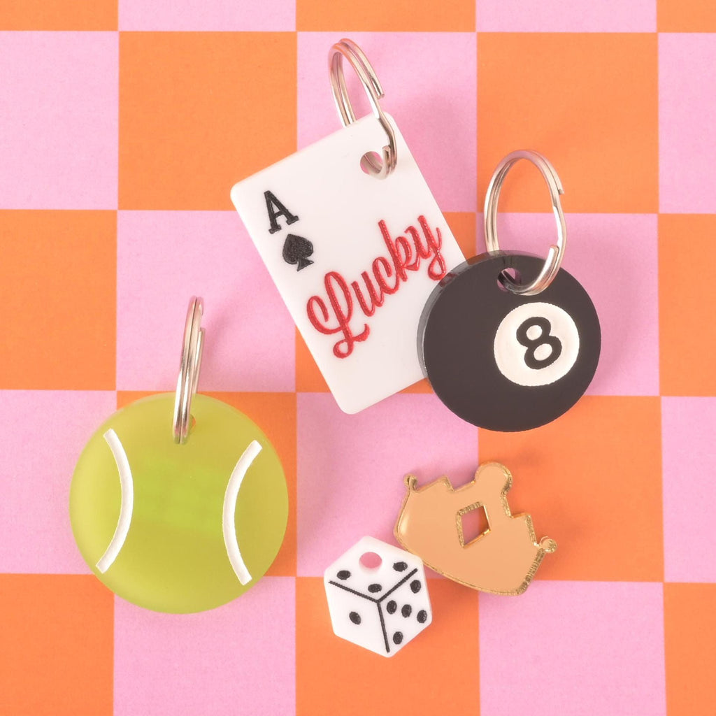 Set of colorful acrylic pet ID tags including a tennis ball, playing card, 8-ball, dice, and crown, shown on a pink and orange checkerboard.