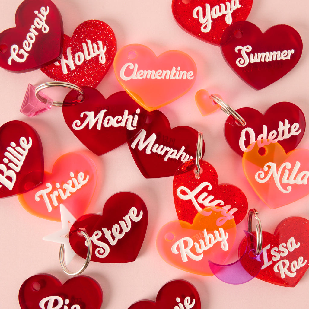 Assorted heart-shaped pet ID tags in red, pink, glitter, and translucent finishes, engraved with pet names like Lucy, Ruby, Clementine, and more.