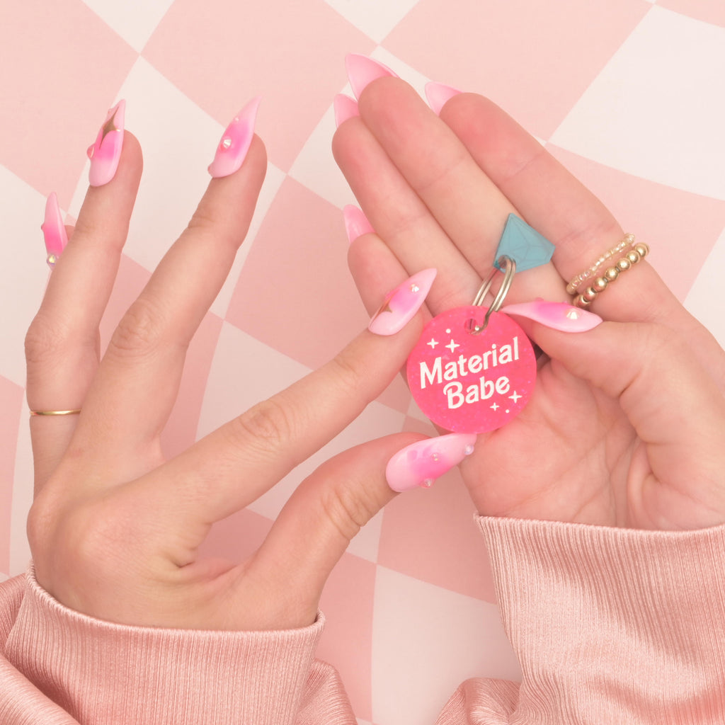 Glitter pink pet tag that reads "material babe" held by handing with pink girly nails on checked pink background