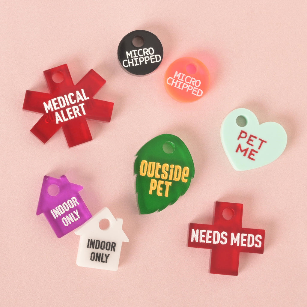 A group of custom, colorful pet ID tags with medical and safety messages. The tags include a red starburst with "MEDICAL ALERT," a black and an orange tag with "MICRO CHIPPED," a green leaf with "OUTSIDE PET," a red cross with "NEEDS MEDS," a mint green heart with "PET ME," and a purple and a white house-shaped tag with "INDOOR ONLY." All are displayed on a solid pink background.
