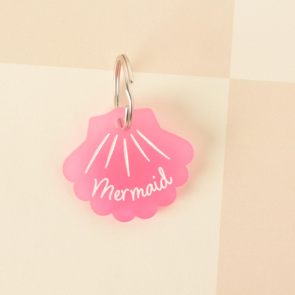 A pink seashell-shaped pet tag with the name "Mermaid" written in white. The tag is attached to a silver ring and is resting on a pink and white checkered background.
