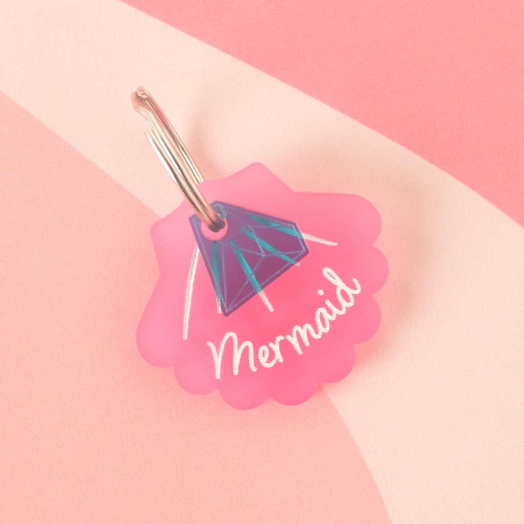 A pink seashell-shaped pet tag with the name "Mermaid" is attached to a translucent blue diamond charm. Both are connected to a silver ring, perfect for a collar. The items are shown on a pink background with a wavy design.