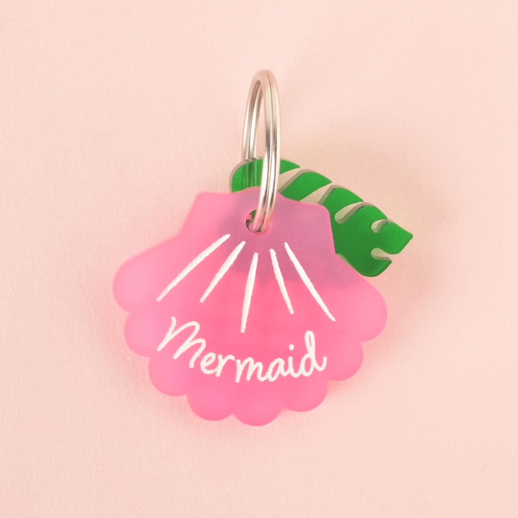 A pink seashell-shaped pet tag with the name "Mermaid" is attached to a green monstera leaf charm. Both are connected to a silver ring, perfect for a collar. The items are shown on a solid pink background.