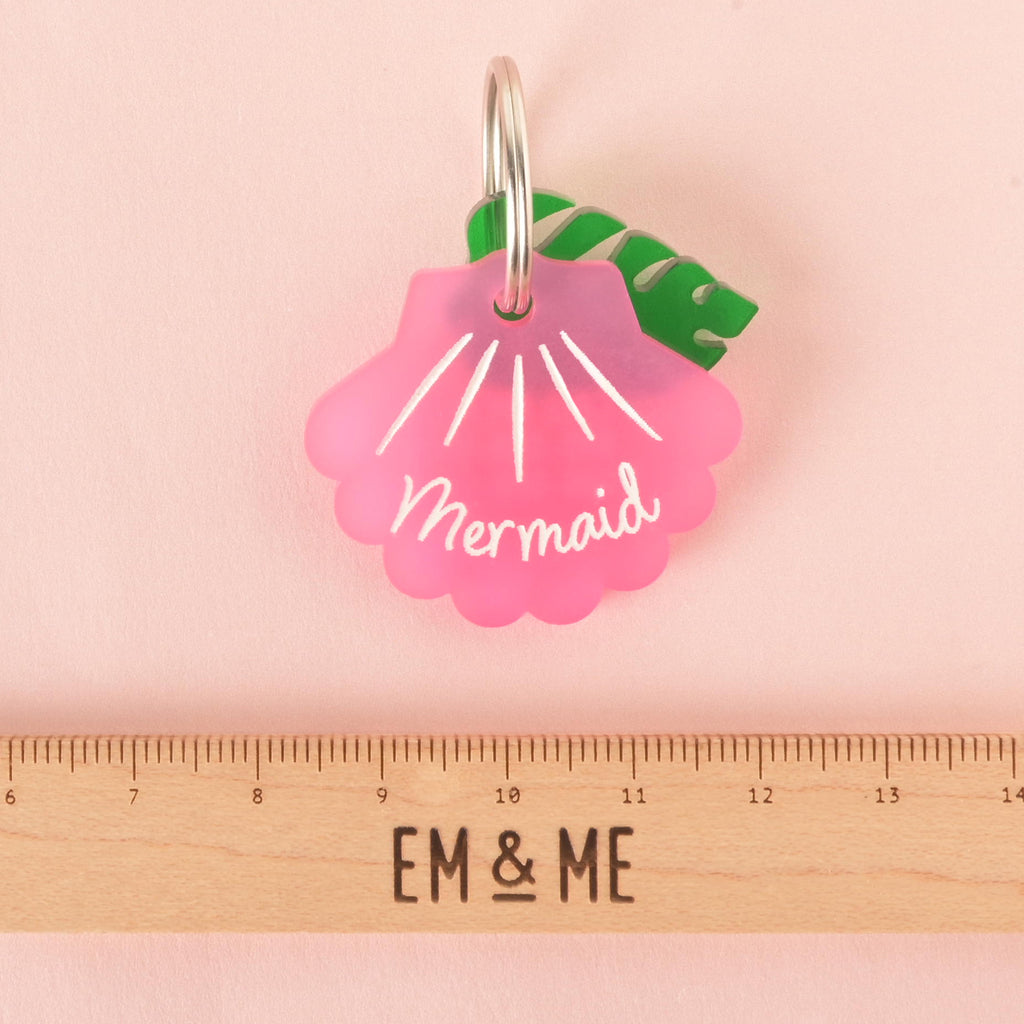 A pink seashell-shaped pet tag with the name "Mermaid" is attached to a green monstera leaf charm. Both are connected to a silver ring, perfect for a collar. A ruler with the "EM & ME" logo rests below the tag, indicating its size. The background is a solid pink color.