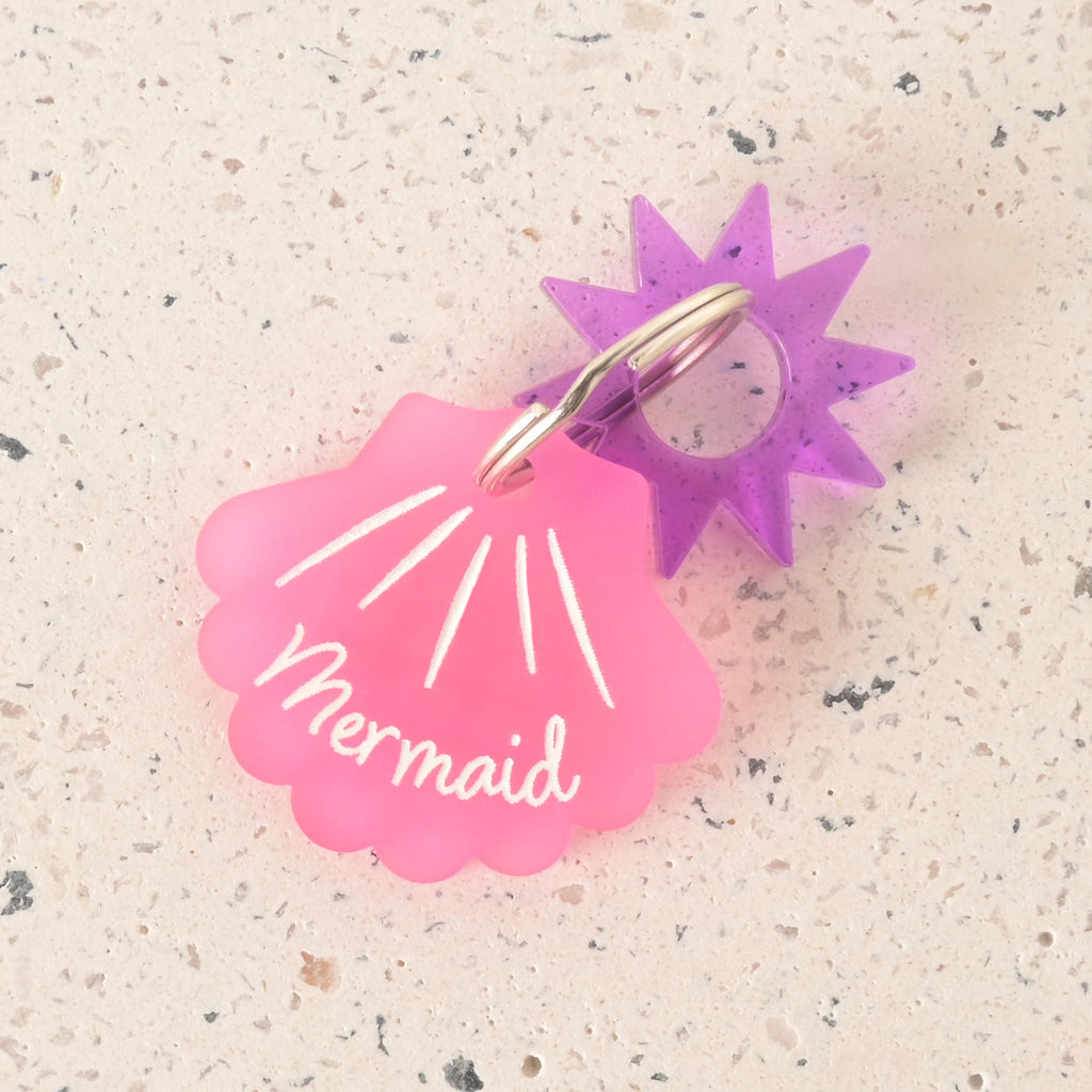 A pink seashell-shaped pet tag with the name "Mermaid" is attached to a purple sun charm. Both are connected to a silver ring, perfect for a collar. The items are shown on a light-colored, speckled surface.