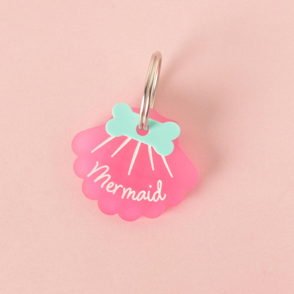 A close-up of a custom, pink, shell-shaped pet ID tag with a shiny silver keyring. The tag is engraved with the word "Mermaid" in white script font and has a light blue bone-shaped charm at the top. The tag is displayed on a solid pink background.