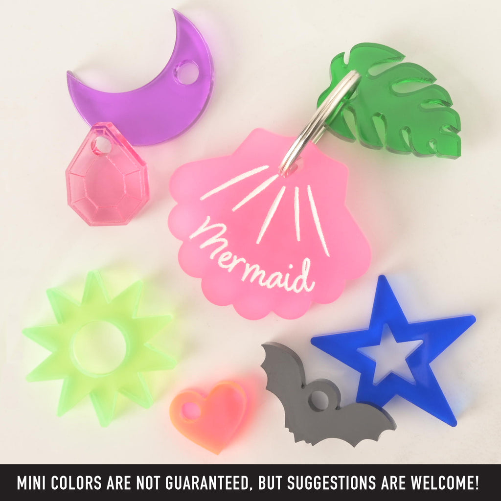 A collection of pet charms and a pet tag. A pink seashell-shaped tag that says "Mermaid" is attached to a green monstera leaf charm. Around them are other charms including a purple moon, a pink gemstone, a neon yellow sun, a neon pink heart, a gray bat, and a blue star. The image also contains the text "MINI COLORS ARE NOT GUARANTEED, BUT SUGGESTIONS ARE WELCOME!" The items are displayed on a white surface.