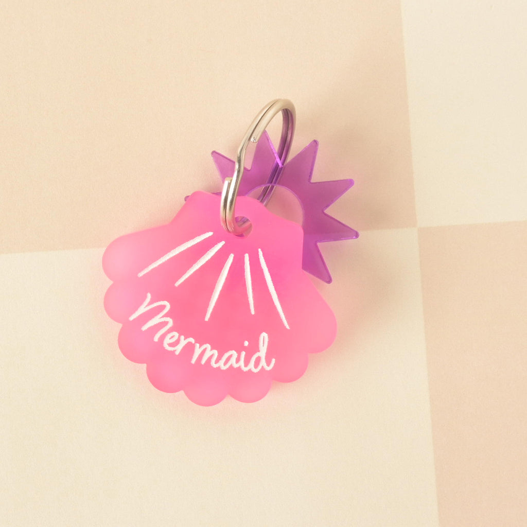  pink, frosted acrylic pet ID tag shaped like a seashell with "Mermaid" engraved on it, paired with a small, clear purple sunburst charm.
