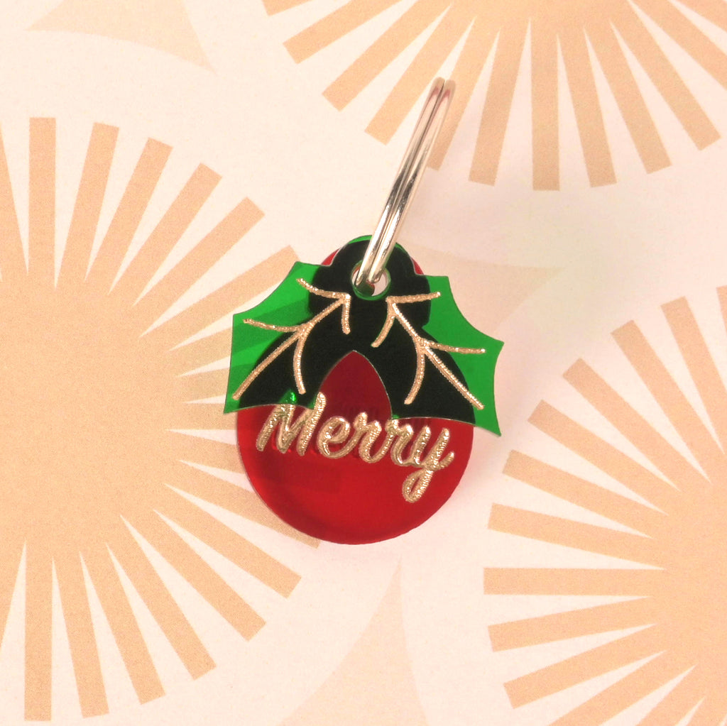 Close-up of a small, red circular pet ID tag with the glitter gold slogan 'Merry.' A green and black acrylic holly leaf charm is layered over the top. The tag is displayed on a retro sunburst patterned background.