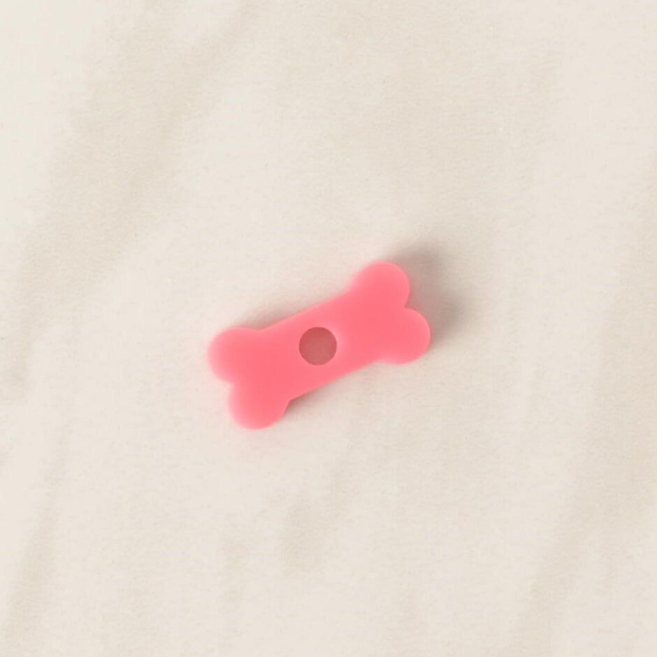 A close-up of a small, translucent pink, bone-shaped pet tag charm. The charm is displayed on a light-colored, speckled countertop or stone surface.