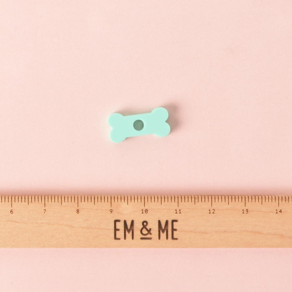 A small, mint green, bone-shaped pet tag charm. The charm is displayed above a wooden ruler with the logo "EM & ME." The background is a solid pink.