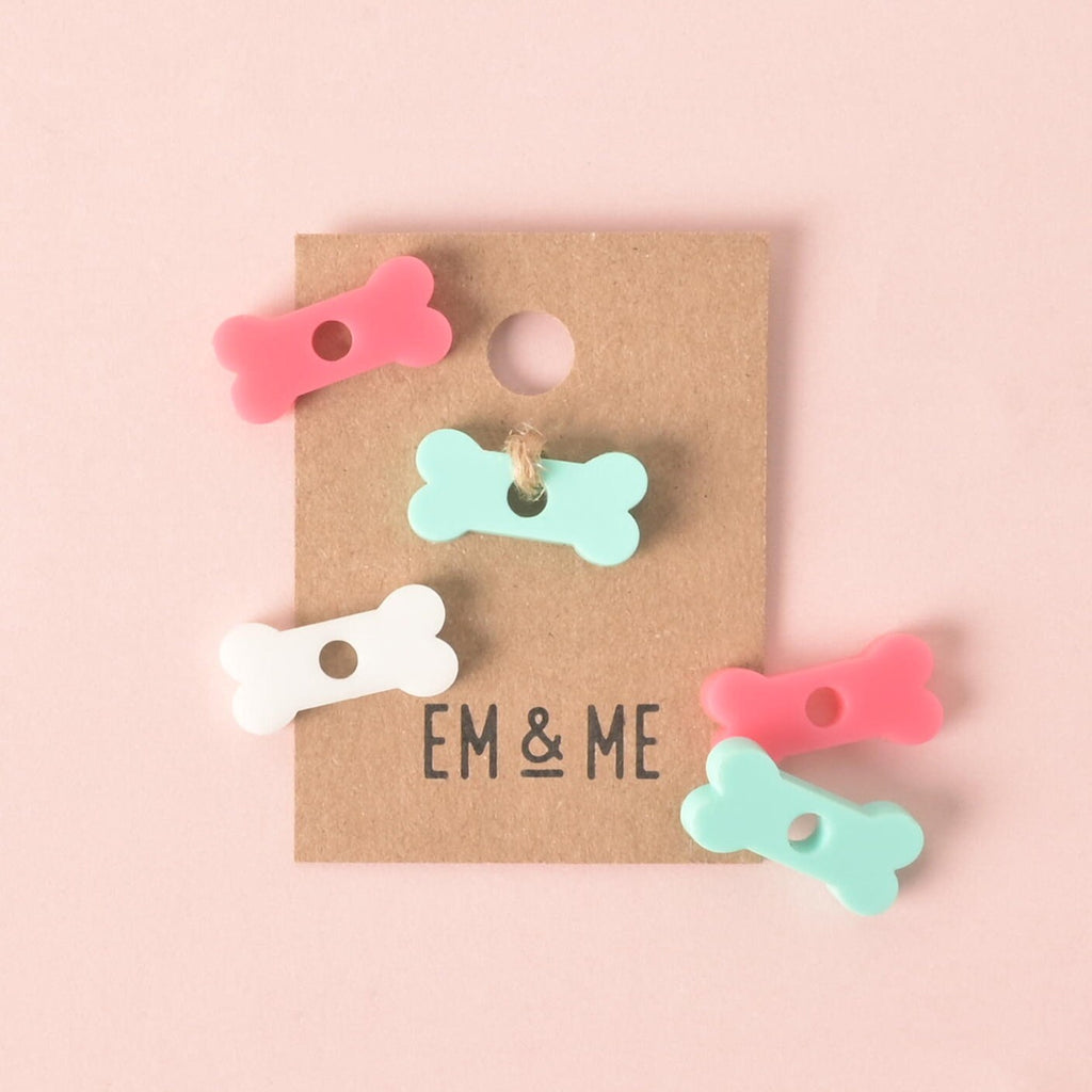 A group of five small, bone-shaped pet tag charms in pastel colors. One mint green bone is attached with a string to a brown cardboard tag that says "EM & ME." Loose charms in pink, white, and mint green are scattered around the tag. The items are displayed on a solid pink background.