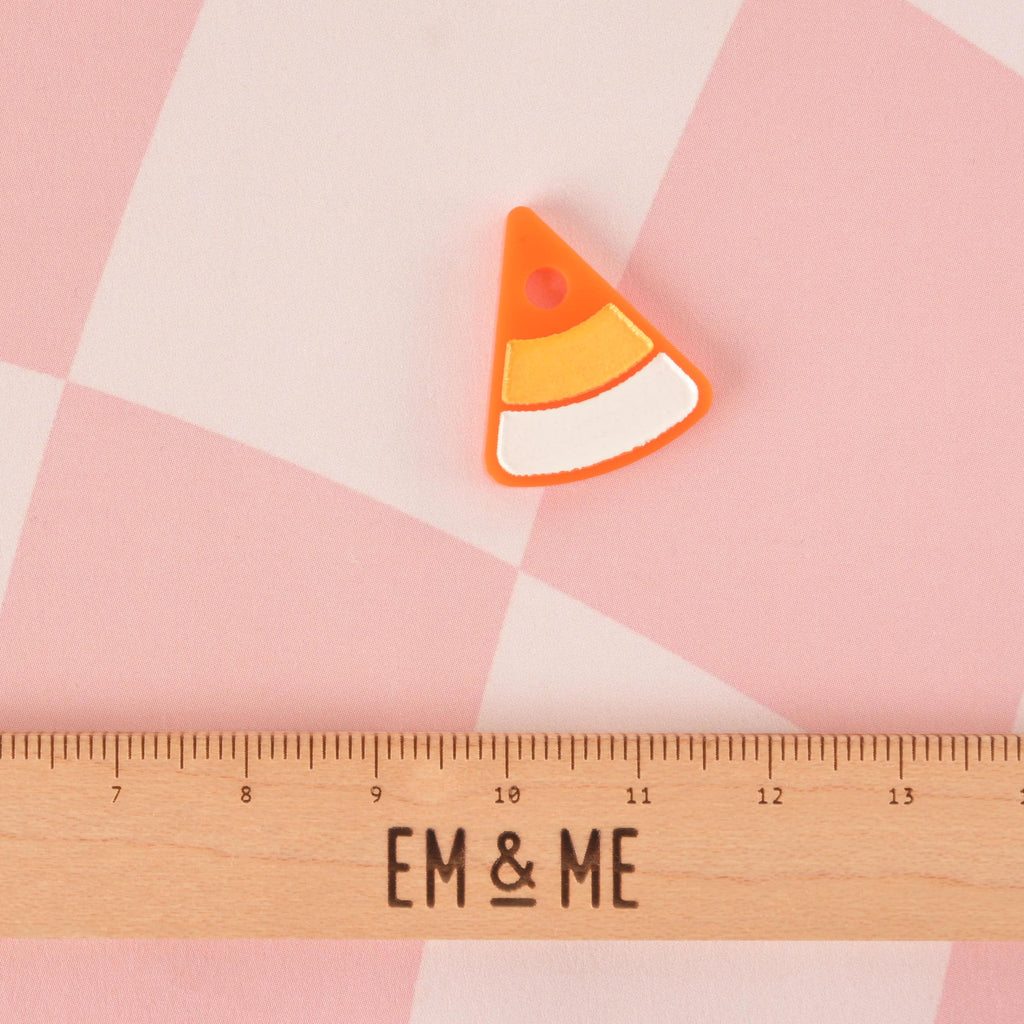 A small, candy corn-shaped pet tag charm. The charm is orange, yellow, and white with a bright orange outline. It is displayed above a wooden ruler with the logo "EM & ME." The background is a pink and white checkered pattern.
