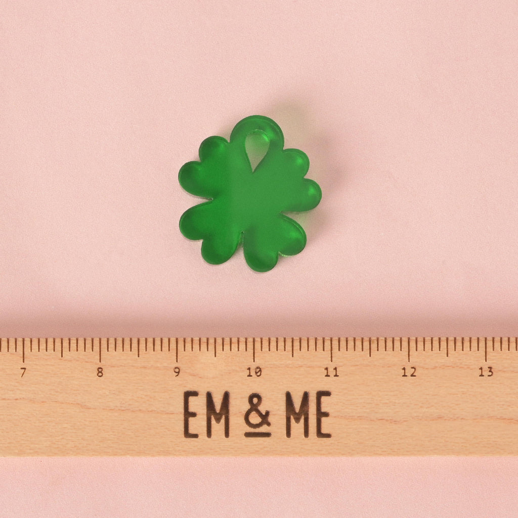 A small, translucent green, four-leaf clover-shaped charm. The charm is displayed above a wooden ruler with the logo "EM & ME." The background is a solid pink.