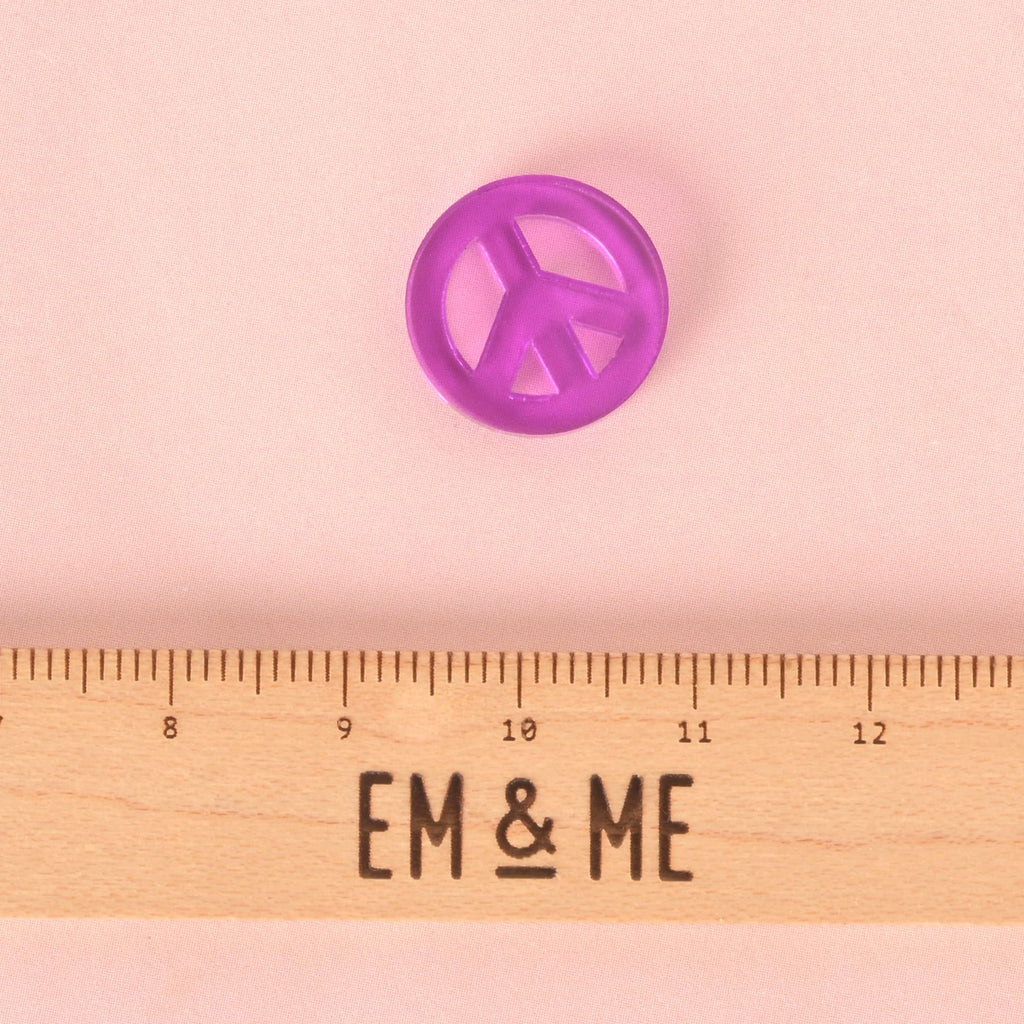 A small, translucent purple peace sign charm. The charm is displayed above a wooden ruler with the logo "EM & ME." The background is a solid pink.